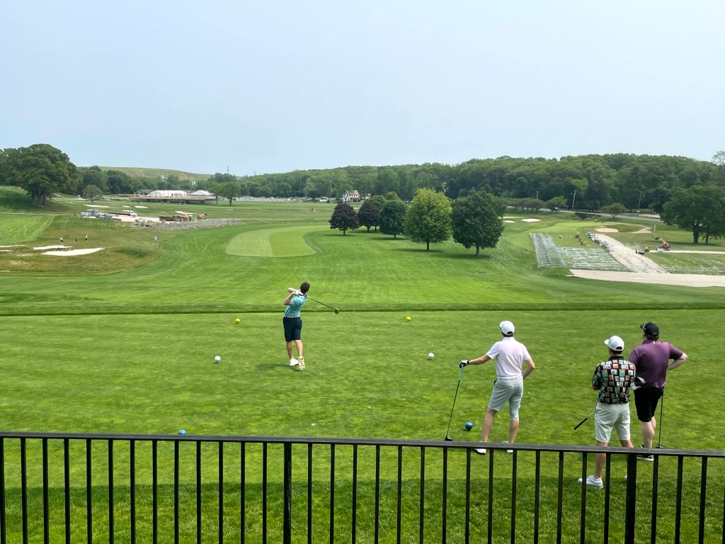 A golfer preparing to tee off at the Black Course in Bethpage State Park, while three spectators watch from a distance.