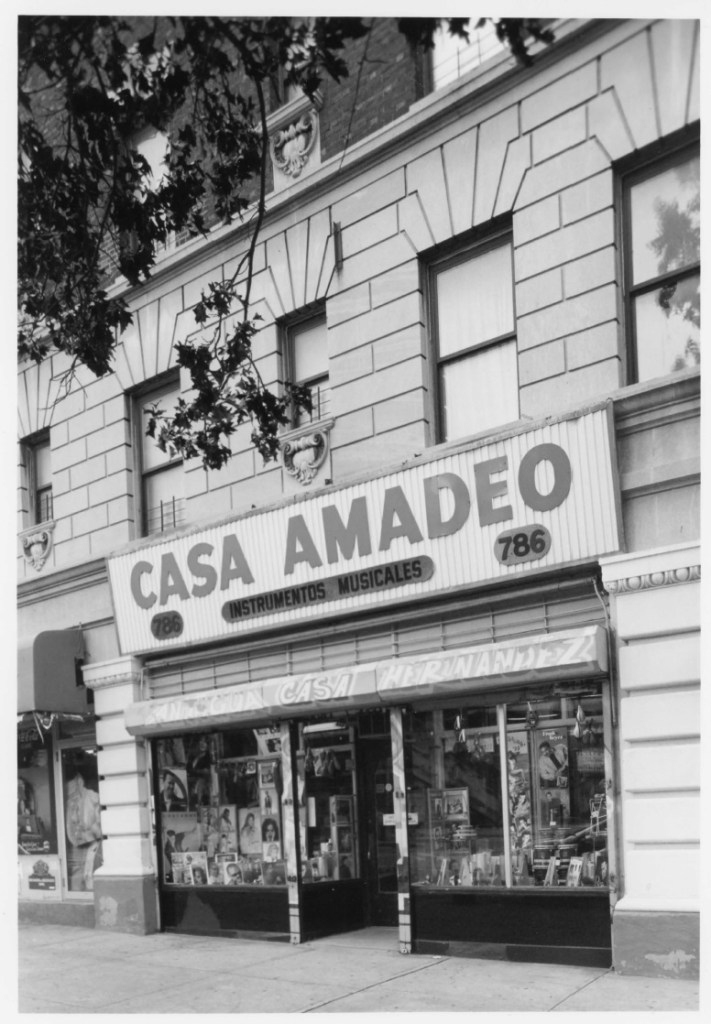 Black and white photograph of Casa Amadeo, a music store located in the Bronx. The storefront features a prominent sign displaying the name 'CASA AMADEO INSTRUMENTOS MUSICALES' along with the address '786' above the entrance. The windows are filled with various music-related items and promotional materials.