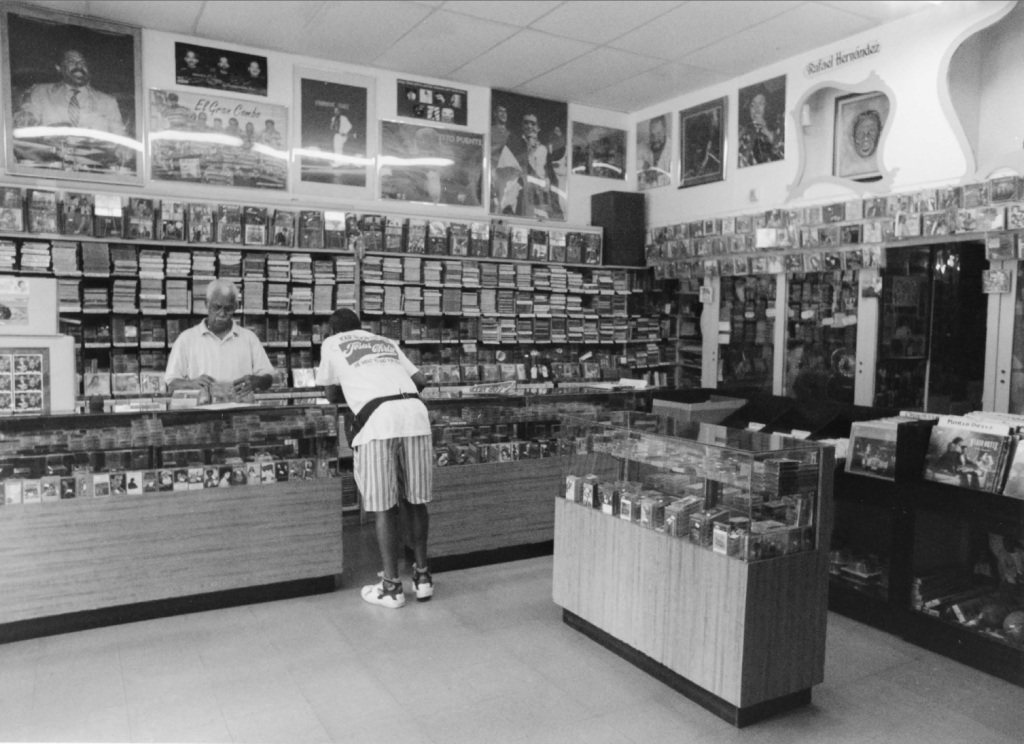 Interior of Casa Amadeo, a Latin music store in the Bronx, featuring shelves filled with music CDs and memorabilia, with a customer and employee engaged in conversation.
