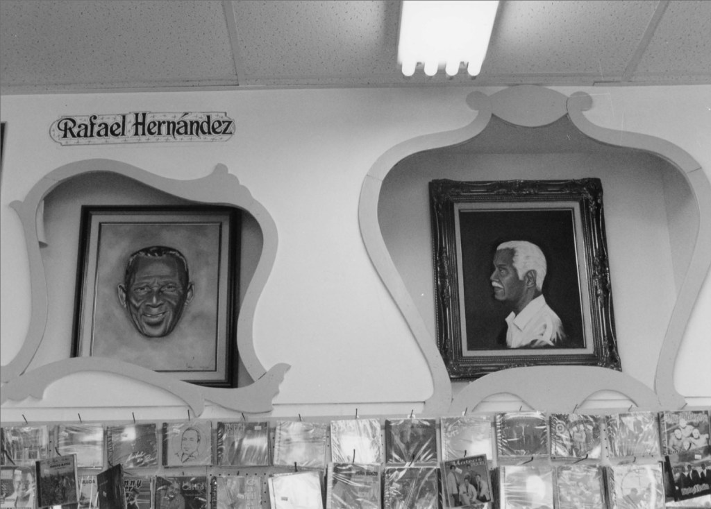 Black and white image featuring two portraits on a wall. The left portrait depicts a smiling man with darker skin and a rounded face, while the right portrait shows a man with lighter skin and a slicked-back hairstyle. Above the portraits is a sign that reads 'Rafael Hernández.' Below the portraits are several music albums displayed on hooks.
