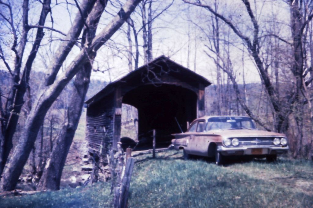A vintage covered bridge nestled among trees, with a classic car parked nearby, set against a pastoral landscape.