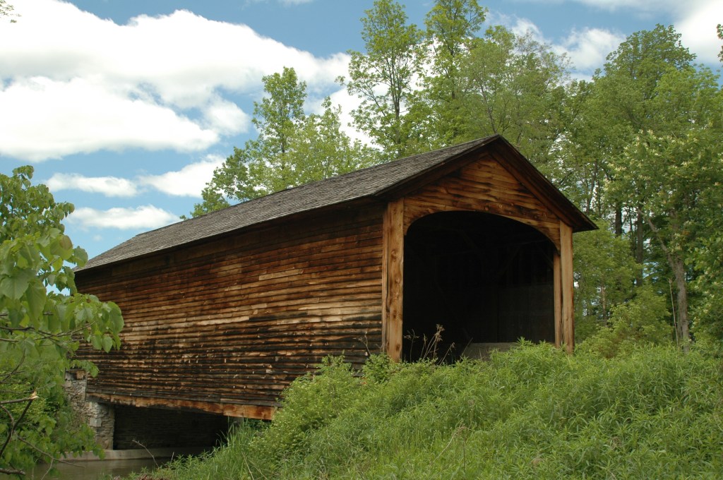 Hyde Hall Covered Bridge in Glimmerglass State Park, a historic wooden structure surrounded by greenery and trees, showcasing its weathered wooden exterior.
