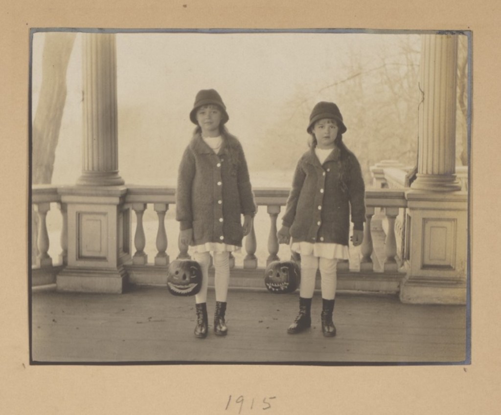 Two young girls stand next to each other on a columned porch. They wear matching hats, sweaters, dresses, and shoes. Both are holding carved pumpkin jack-o-lanterns.