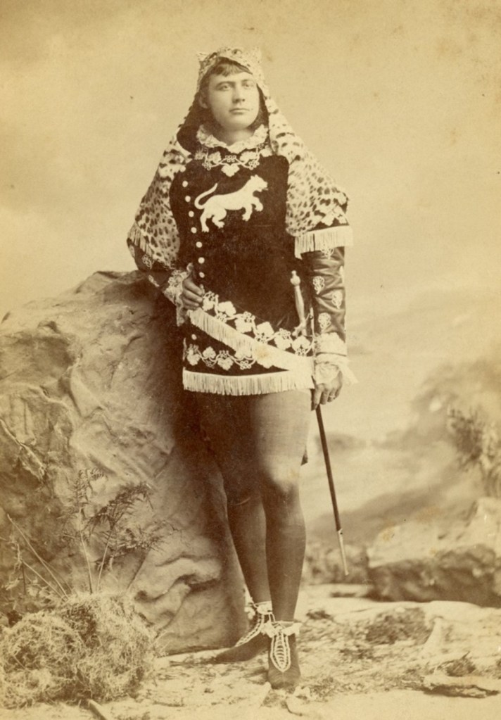 Young man dressed in a short, hooded medieval tunic decorated with lion emblem and fringe; tights; matching shoes; and a sword. He is posed in a photography studio, standing on sandy ground and next to a large boulder.
