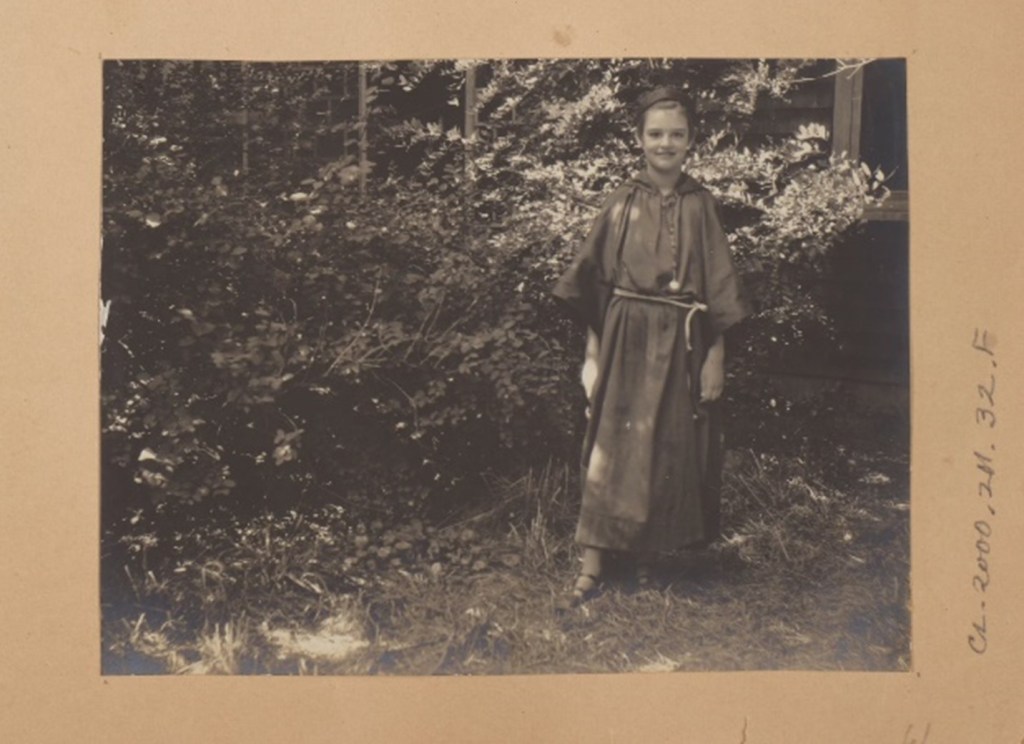 A child stands outside and is posed for the camera. She is wearing long, oversized friar robe with a rope belt, hat, and sandals and is smiling at the camera