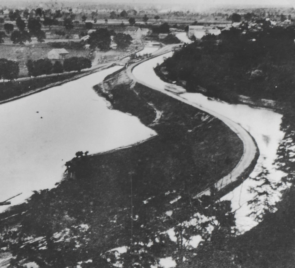 Black and white historical photograph of a winding canal with trees and buildings in the background, illustrating the early infrastructure of the area.