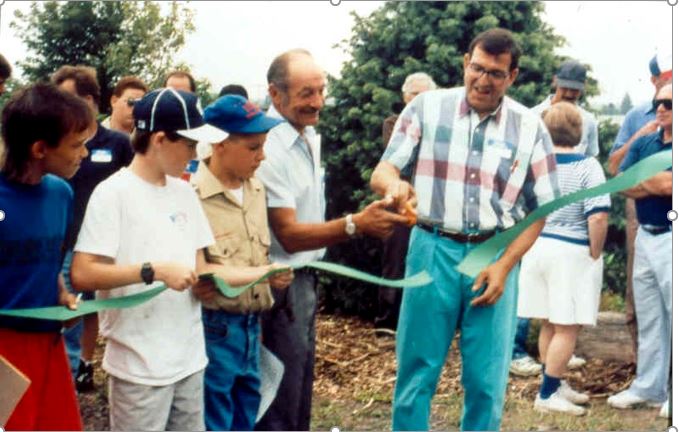 A group of people, including children and adults, gather to cut a green ribbon during a ceremonial event, signifying the opening of a new park or community space.