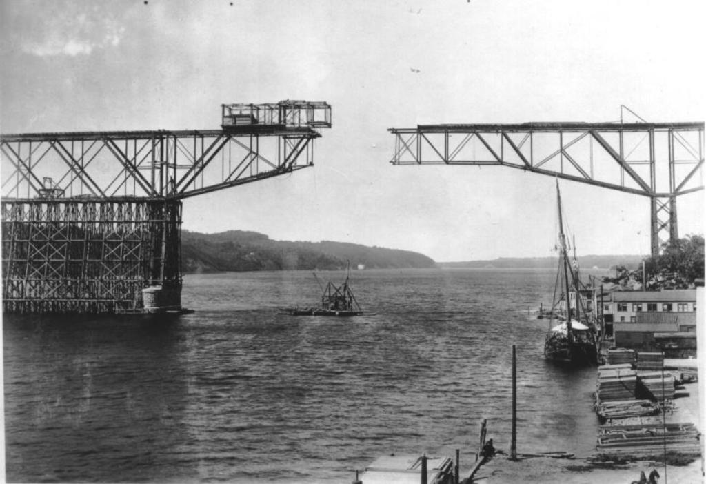 Historic image of the construction of a bridge over a river, featuring large metal structures and scaffolding near the water.