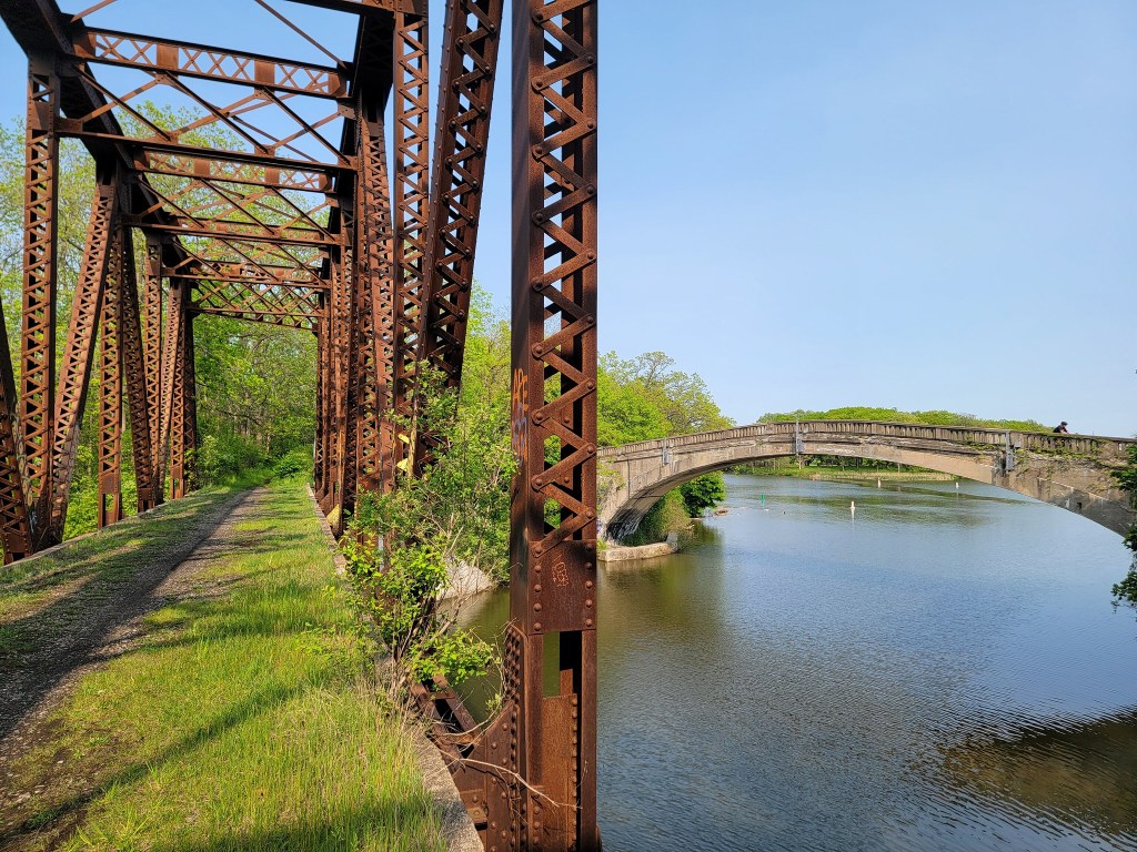 View of an old, rusty railway bridge alongside a green, overgrown path and a modern concrete bridge crossing a calm river on a clear day.