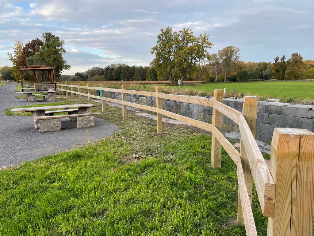 A wooden fence and picnic tables along a walking path with grassy areas and trees in the background at a state park.