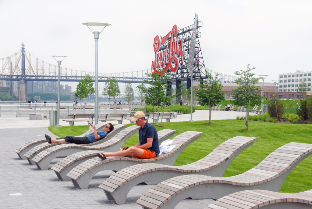 People relaxing on modern benches at Gantry Plaza State Park with the iconic Coca-Cola sign and the Queensboro Bridge in the background.