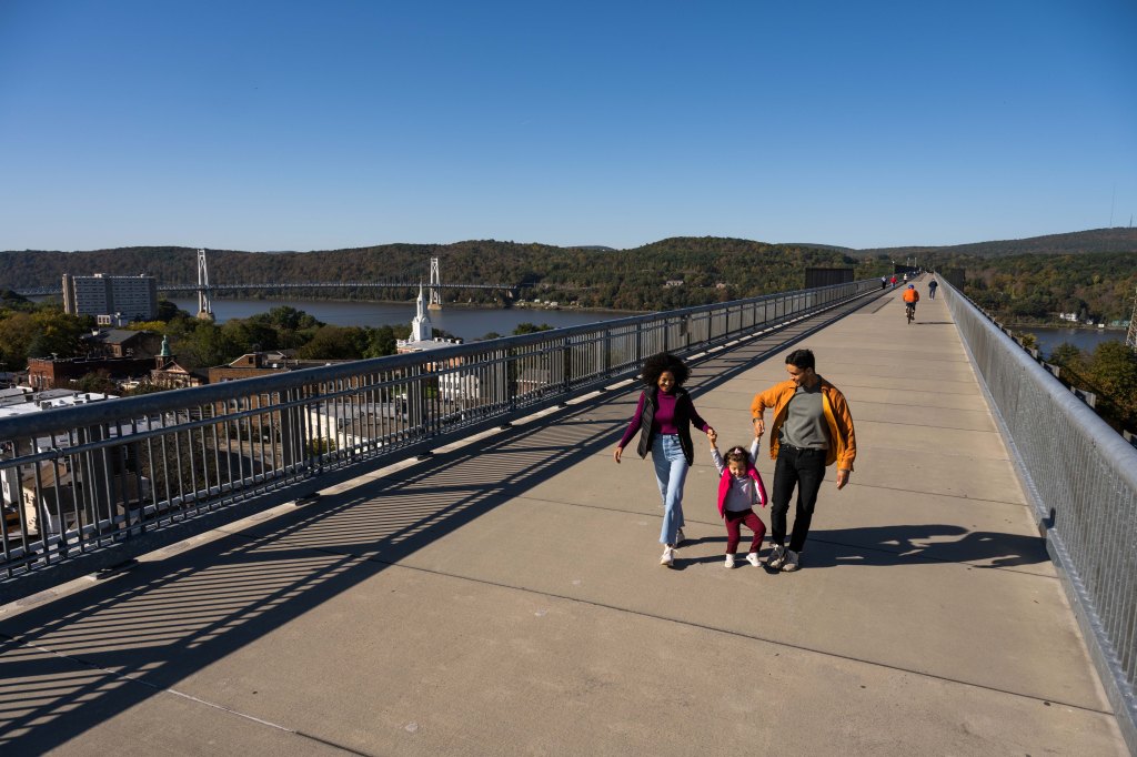 A family with a child walking hand-in-hand along the Walkway Over the Hudson, an elevated pedestrian bridge, with scenic views of the river and surrounding hills in the background.