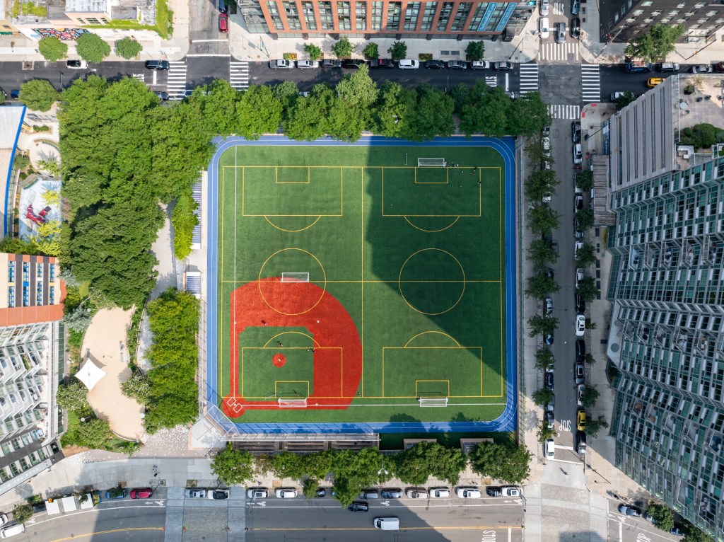 Aerial view of a multi-use sports field in an urban area, featuring a soccer pitch with yellow lines and a baseball diamond in red, surrounded by trees and residential buildings.