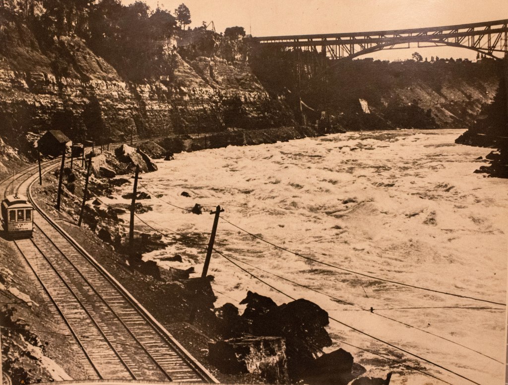 Historic black and white image of a trolley car traveling along railway tracks beside the raging waters of the Niagara Gorge, with a bridge visible above.