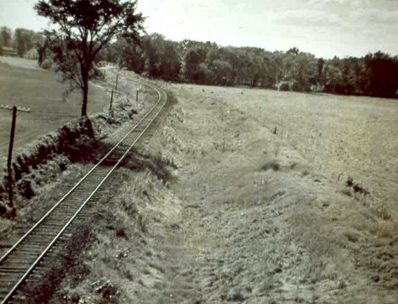 A historical photograph of a curved railroad track running through a rural landscape, with a tree on the left and open fields stretching out in the background.