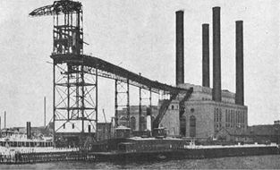 Historical photograph of an industrial structure with large smokestacks, a loading dock, and a barge on the water, showcasing early 20th-century infrastructure.
