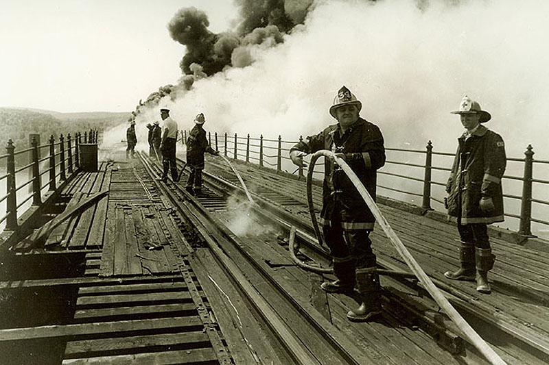 Historical black and white photo of firefighters battling a fire on a wooden bridge. Smoke billows in the background as firemen work with hoses.