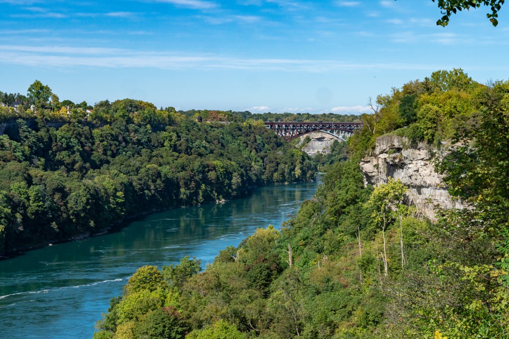 A scenic view of the Niagara Gorge, featuring lush green trees and cliffs alongside the flowing river, with an old railway bridge visible in the background.