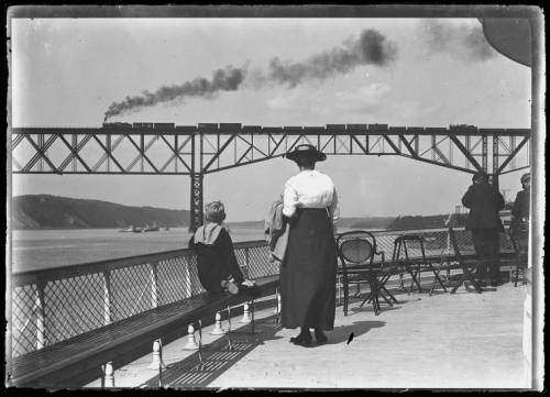 A historical black and white photograph depicting a group of people on a waterfront deck, with a train crossing an iron bridge in the background, emitting smoke over the river.