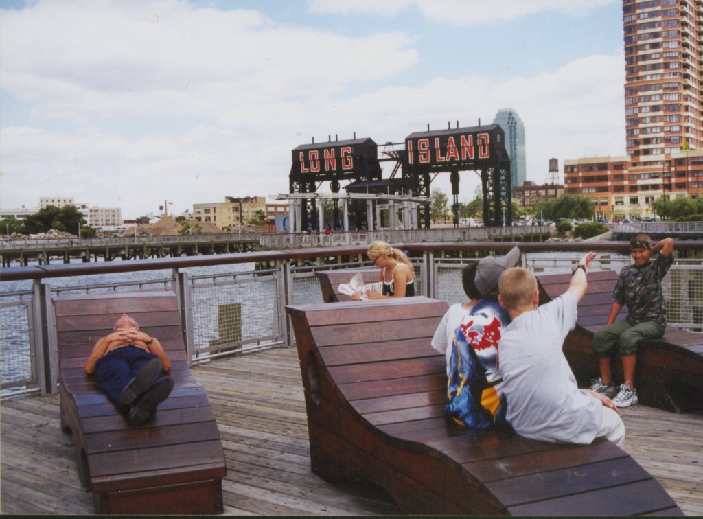 Visitors relax on wooden lounge chairs at Gantry Plaza State Park, with the iconic 'LONG ISLAND' sign and the East River in the background.