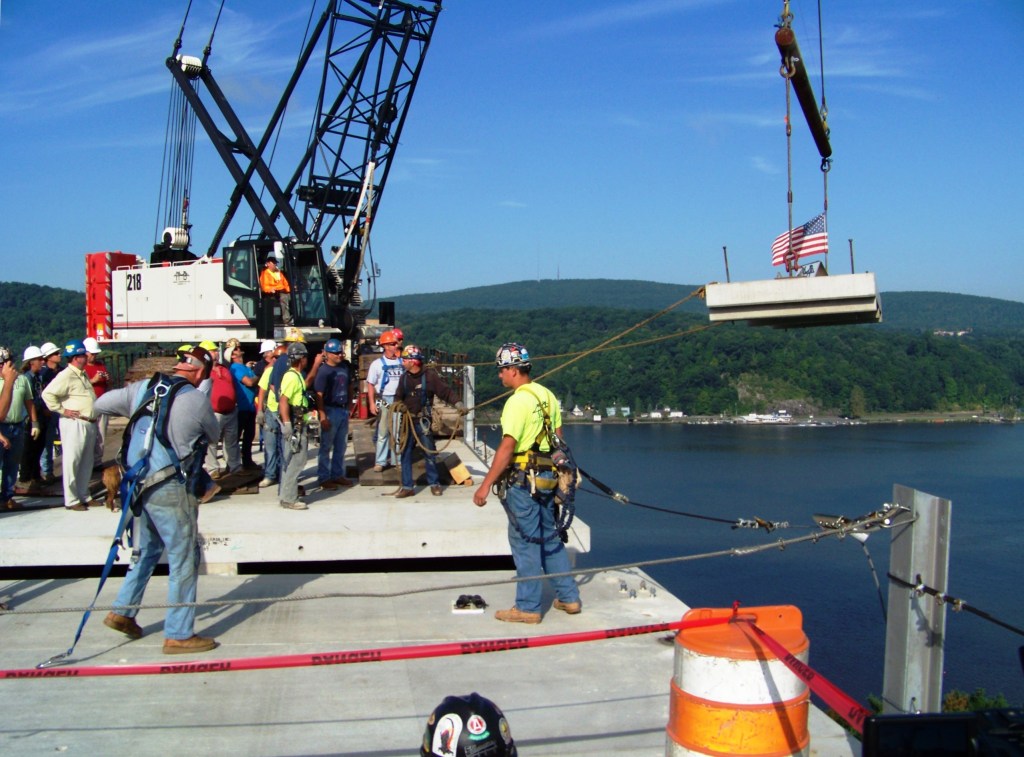 Construction workers and a crane are seen on a bridge site, with a flag attached to a lifting platform and a scenic view of the water and hills in the background.