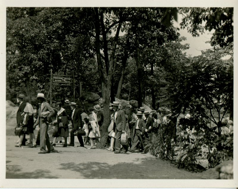 A historical black and white photograph showing a group of people, including adults and children, walking along a path in a forested area, dressed in early 20th-century attire, with some carrying bags.