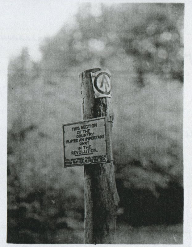 Black and white photograph of a wooden post with a sign indicating the historical significance of the area during the revolution, featuring a peace symbol and text about its importance.
