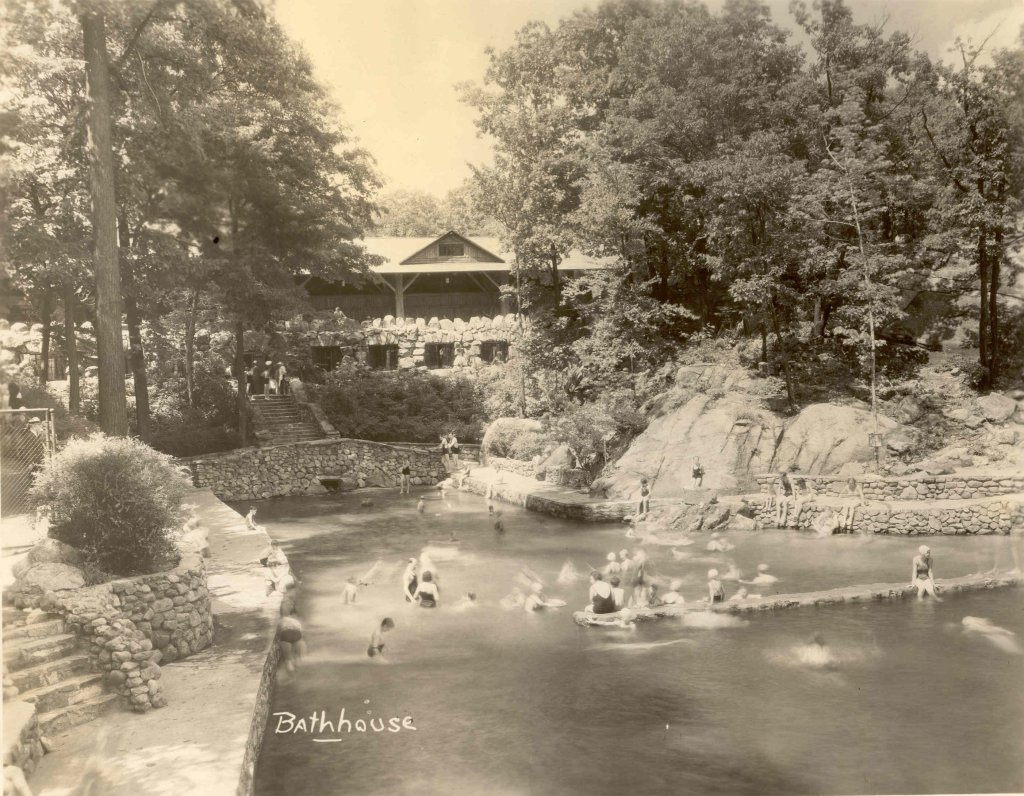 Historic black and white image of a bathhouse and swimming area surrounded by trees, with people swimming and relaxing in the water.