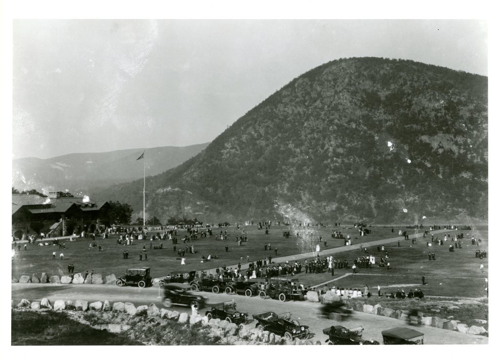 Historic black and white photograph of a large crowd gathered on the lawn by Bear Mountain State Park, with cars parked along a road and a mountain in the background.