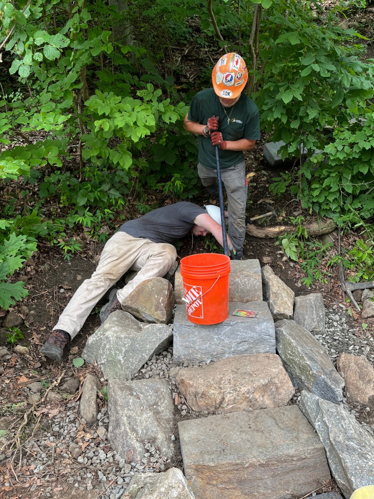 Two trail crew members working on trail maintenance, laying stones to create a stable pathway in a wooded area.