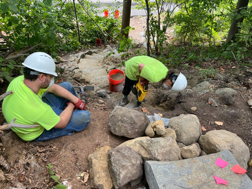 Two trail crew members working on a stone step construction project in a forested area, surrounded by rocks and greenery.