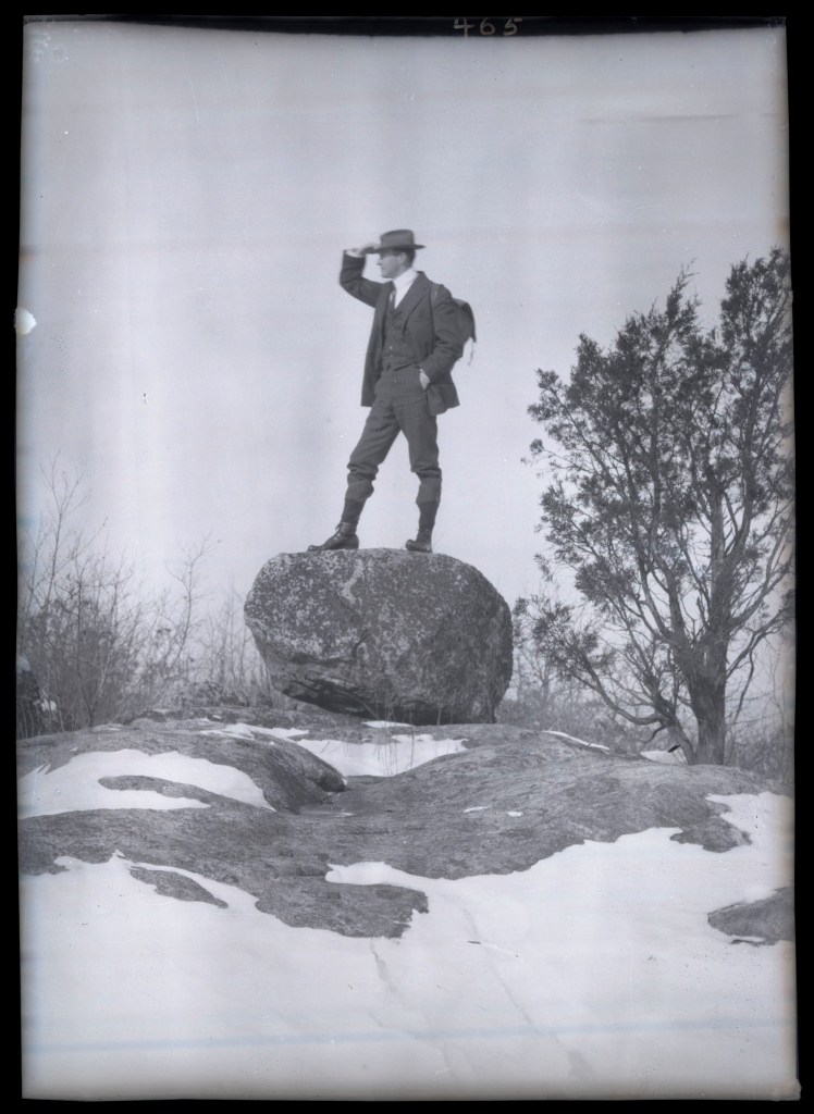 A vintage black and white photograph of a man in formal attire standing confidently on a large rock, looking into the distance while shielding his eyes from the sun. The surrounding landscape is rocky and barren, with patches of snow visible.
