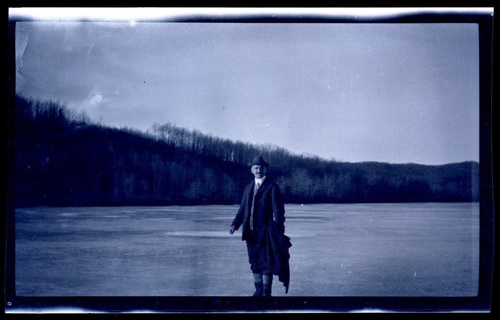 A historical black and white photograph of a man standing on a frozen lake, wearing a suit and a hat, with trees in the background.